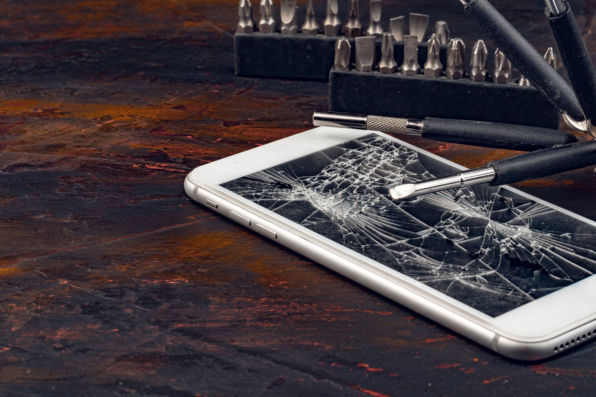 Broken smartphone with shattered screen and screwdriver on wooden surface.